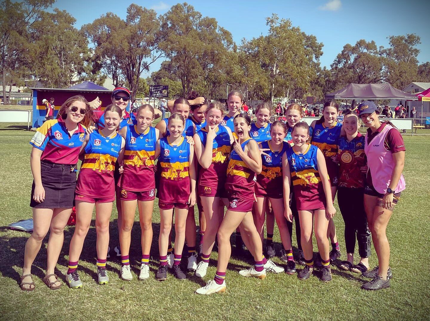 A group of girls footballers in arms on the field after winning a game. They are wearing maroon, yellow and blue uniforms. 
