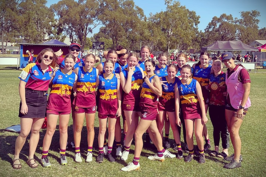 A group of girls footballers in arms on the field after winning a game. They are wearing maroon, yellow and blue uniforms. 