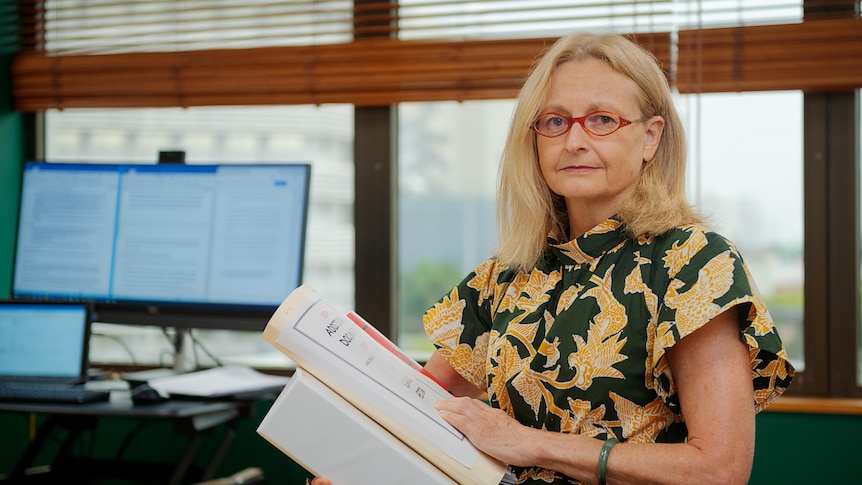 A woman standing inside an office and looking serious while holding several thick documents. 