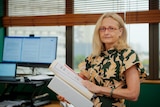 A woman standing inside an office and looking serious while holding several thick documents. 