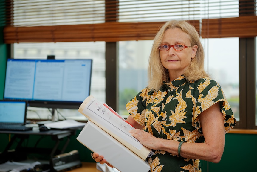 A woman standing inside an office and looking serious while holding several thick documents. 