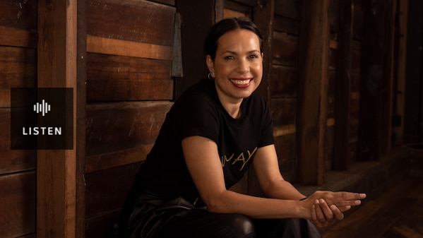 A woman with black hair pulled back wears a black shirt that reads 'Always' in gold and sits against a wooden wall. Has Audio.