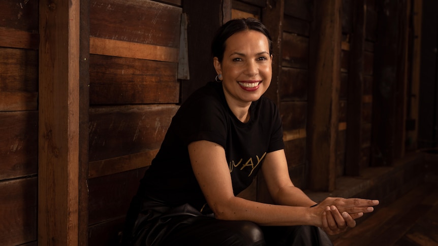 A woman with black hair pulled back wears a black shirt that reads 'Always' in gold and sits against a wooden wall.