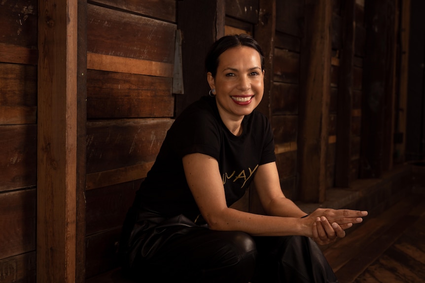 A Kokatha woman with black hair pulled back wears a black shirt that reads 'Always' in gold and sits against wooden wall
