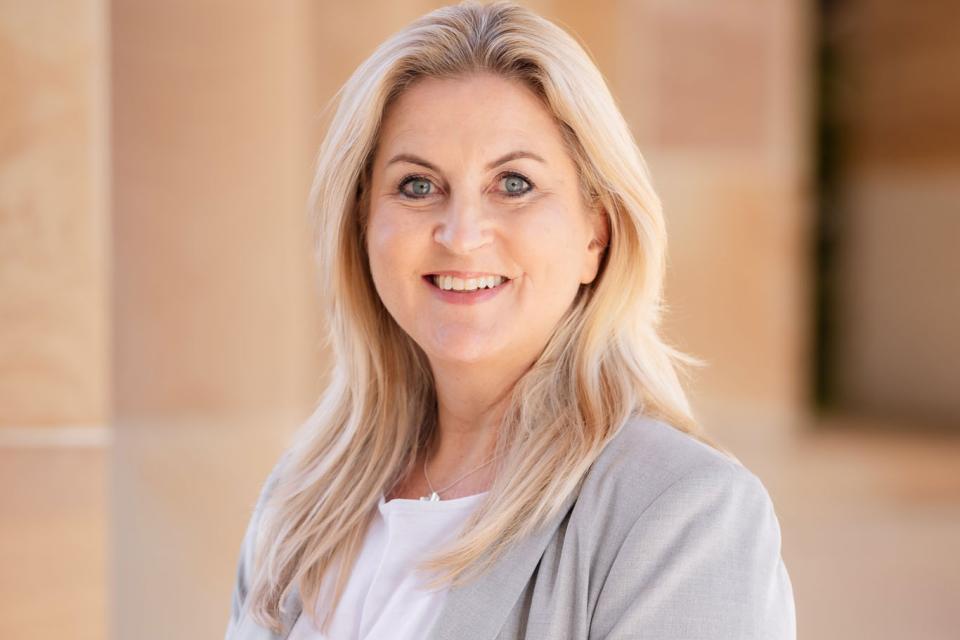 A woman with long blonde hair in a light grey jacket with a white top stands in front of a sandstone building.