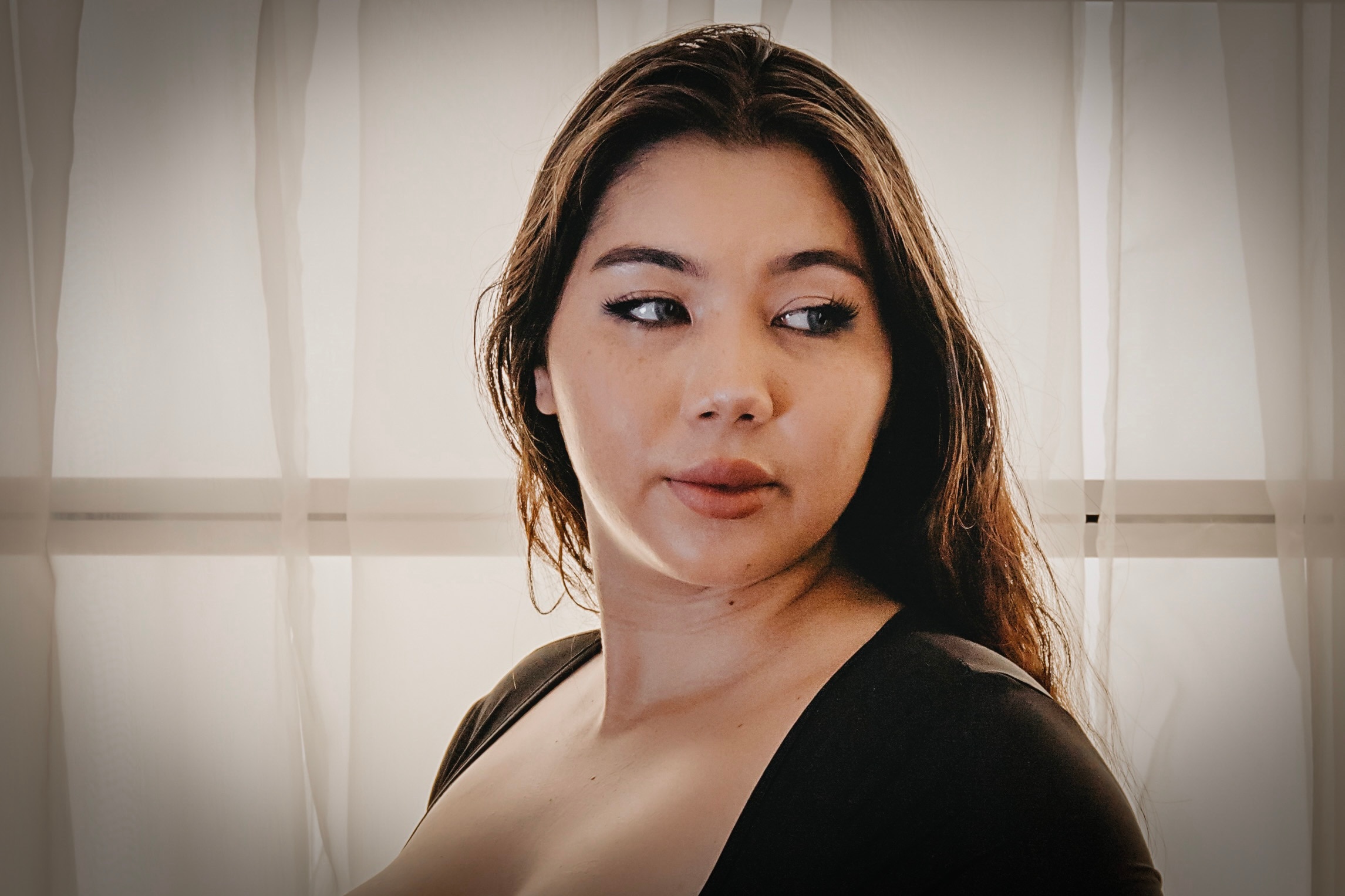 A woman peers to the side in front of a window wearing a black shirt