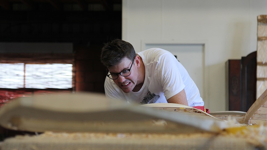 A man pulling a concentrated face while working on a wooden surfboard.
