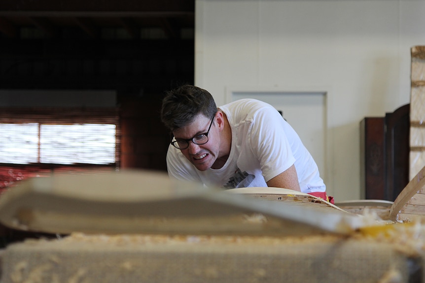 A man pulling a concentrated face while working on a wooden surfboard.