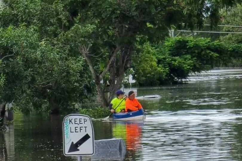 Two people wearing high-vis navigate a flooded street in a kayak.