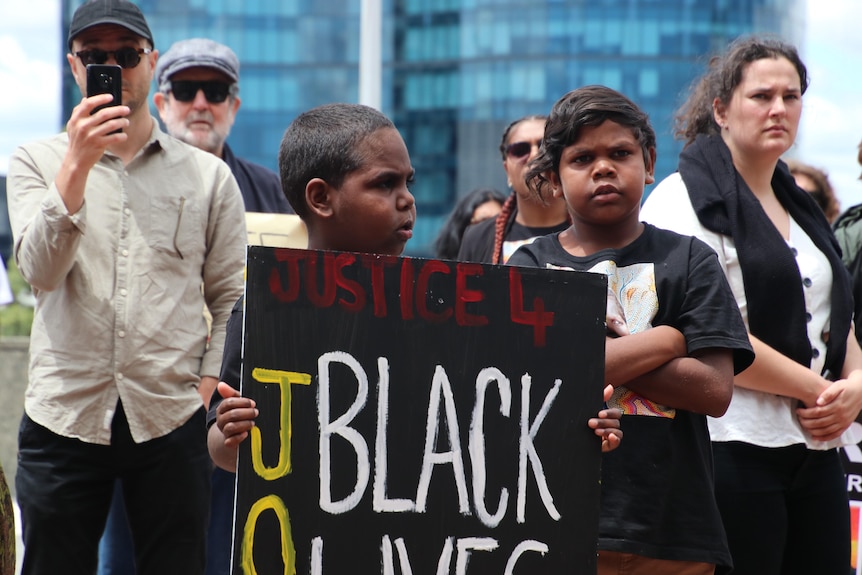 Caesar holds a sign up, another little boy stands next to him with his arms crossed.