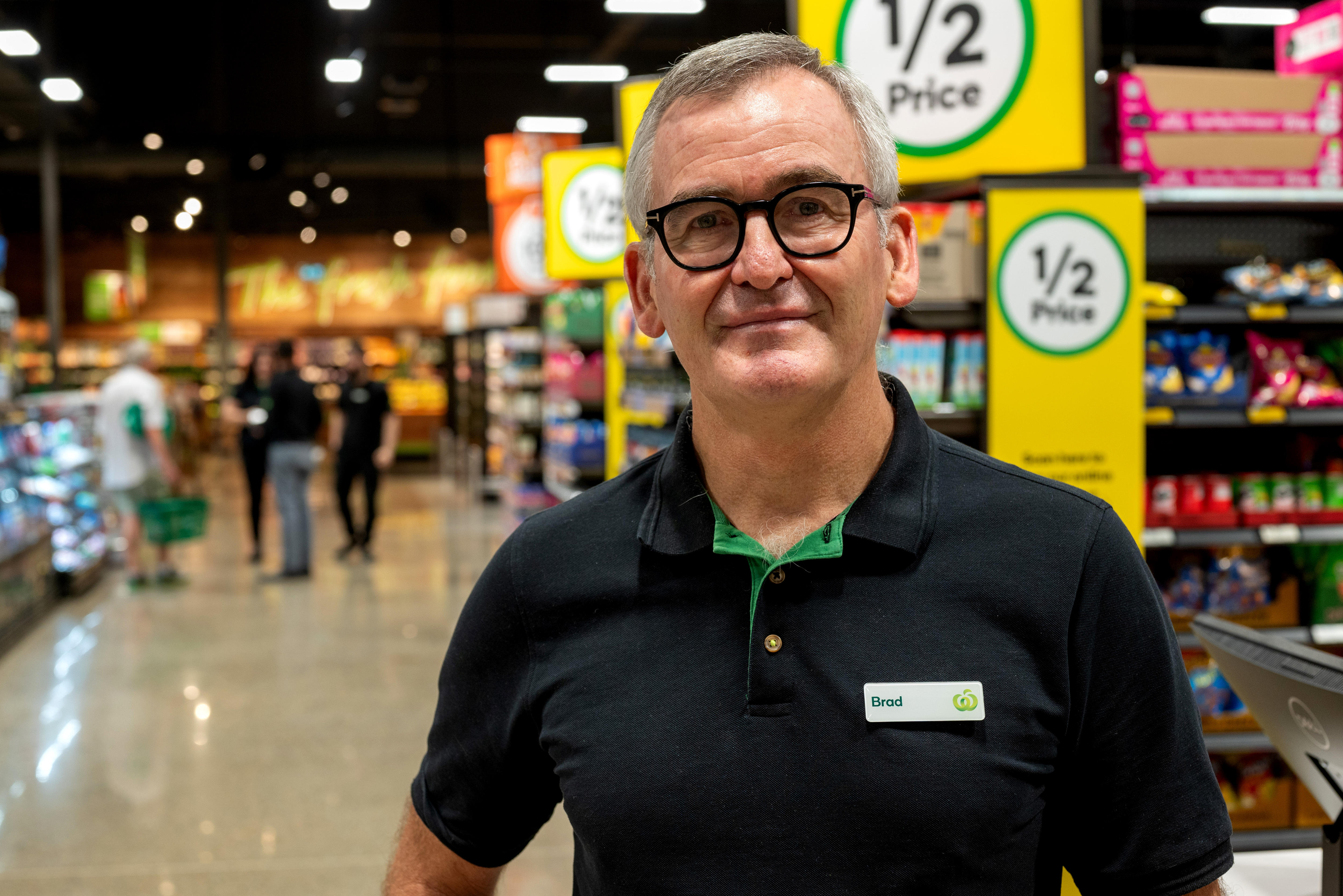 A man wearing a Woolworths polo shirt with the name tag 'Brad' stands in a supermarket.