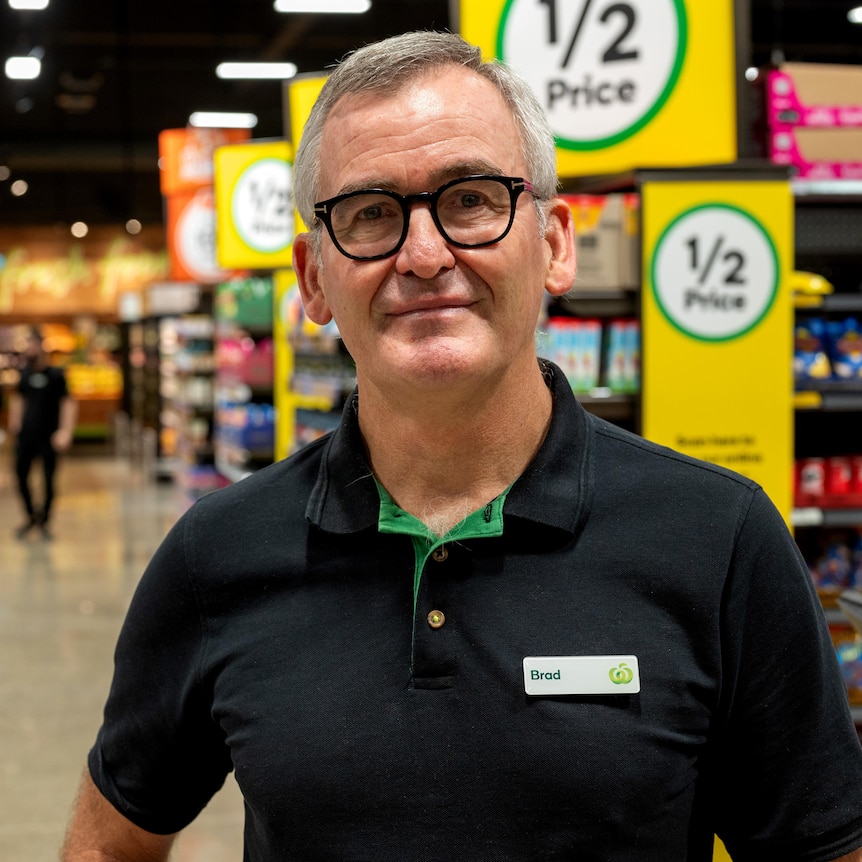 A man wearing a Woolworths polo shirt with the name tag 'Brad' stands in a supermarket.
