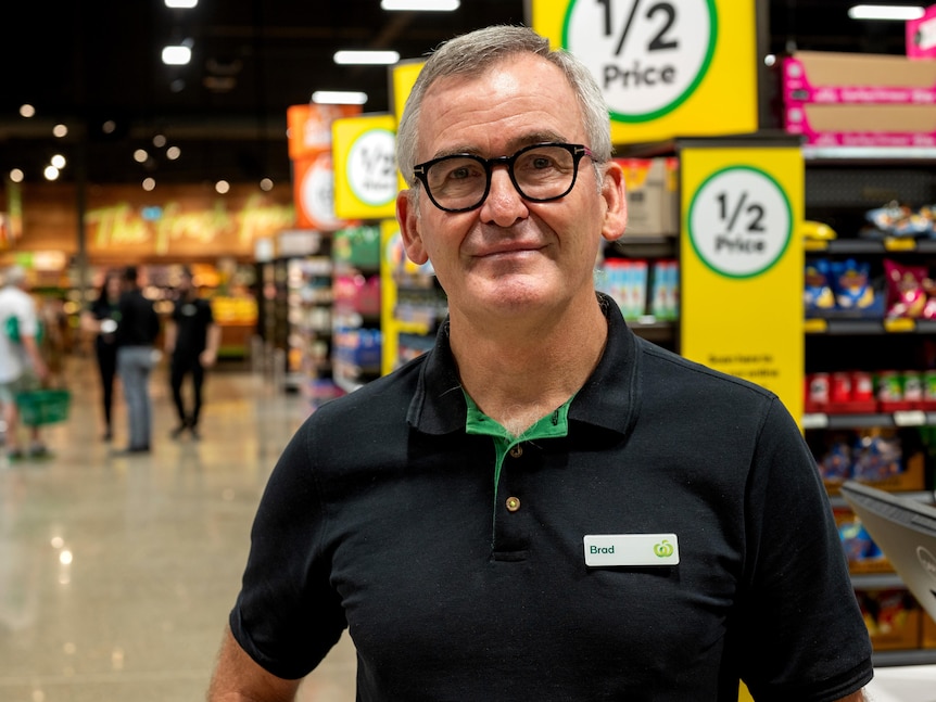 A man wearing a Woolworths polo shirt with the name tag 'Brad' stands in a supermarket.
