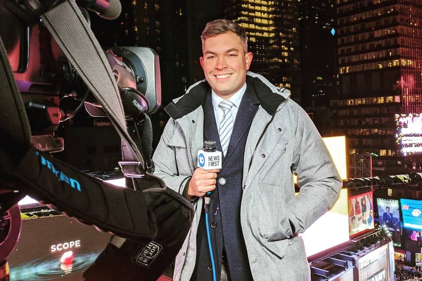 Man holding a microphone and standing in front of a TV news camera. 