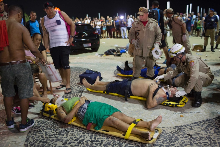 Firefighters give first aid to injured people on the beach. There are three people lined up on stretchers wearing neck braces.