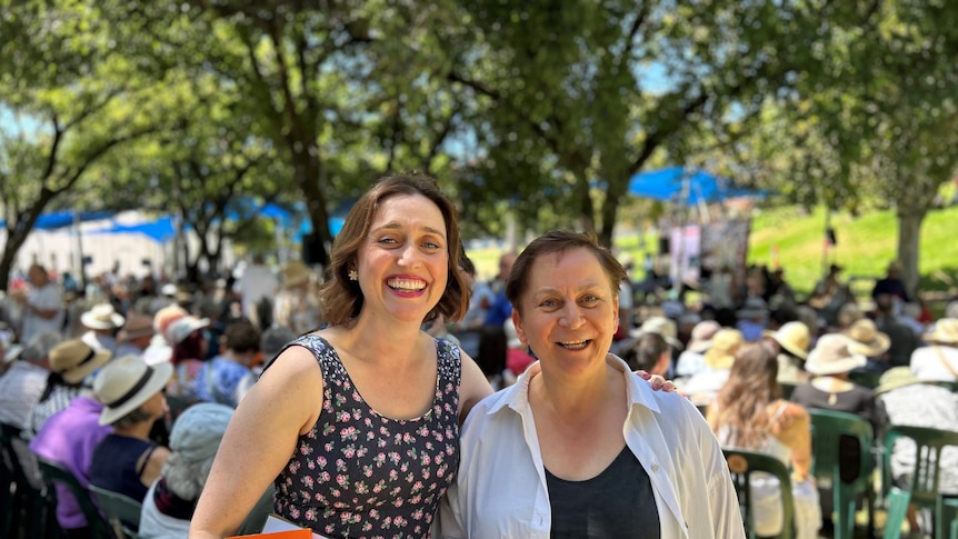 Two women smiling and standing next to eachother outside with festival audience sitting under trees in the background 