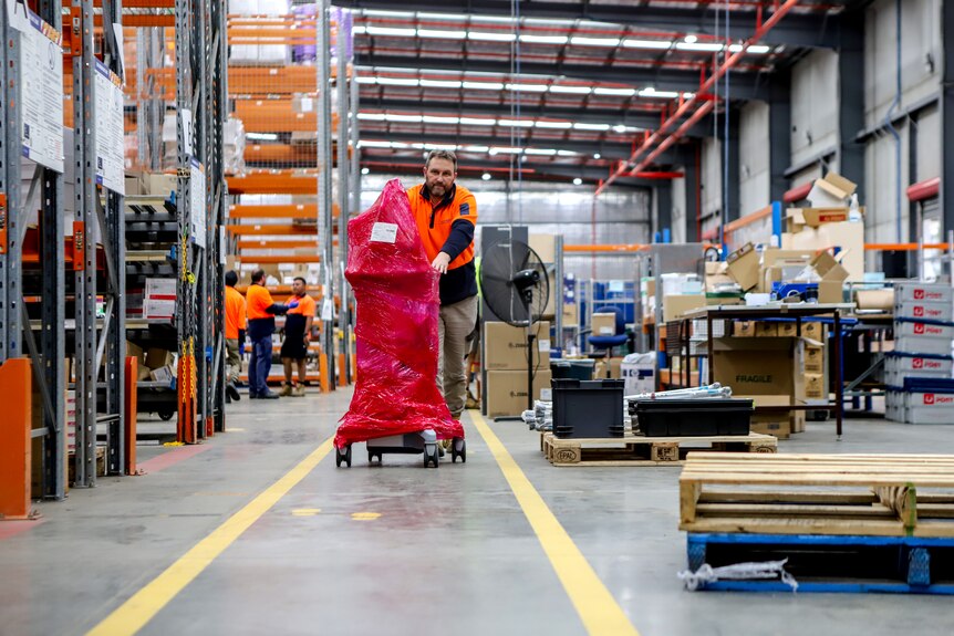 Man in high-vis shirt wheels red plastic wrapped ventilator across warehouse floor next to  large shelves