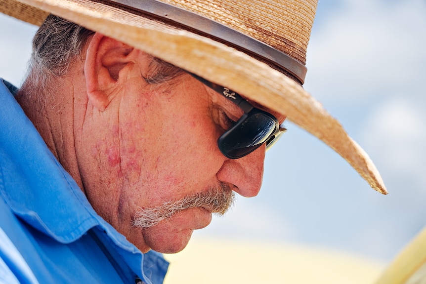 Profile of an older man wearing a hat, sunglasses and a blue shirt.