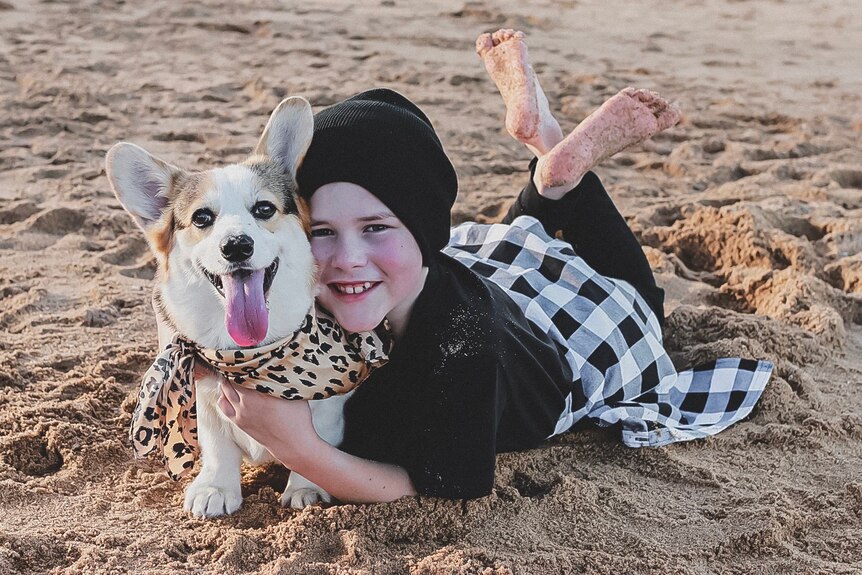 A young boy lies on the beach dressed in black with a white and tan corgi.