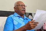 Sitiveni Rabuka wearing a blue shirt speaks at a table holding a piece of paper