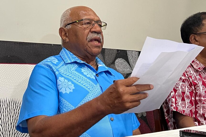 Sitiveni Rabuka wearing a blue shirt speaks at a table holding a piece of paper