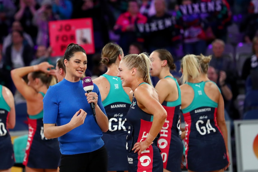 A dark-haired interviewer in a royal blue skivvy speaks to a netballer after a game and there are other players behind them