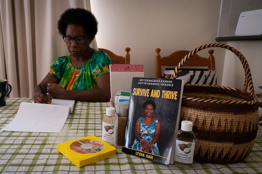 PNG woman sits at table writing with book, basket and CDs in foreground