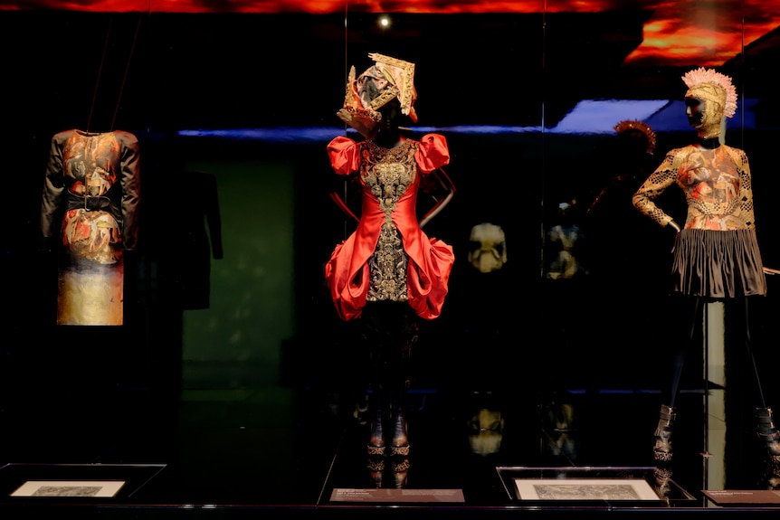 Three mannequins dressed in elaborate and intricate dresses stand on a black podium under bright orange lights in a gallery.