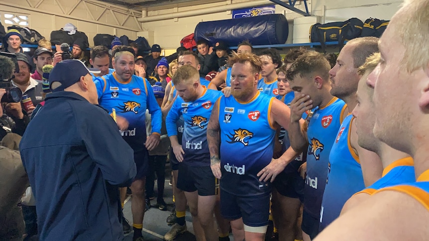 A man in a dark blue coat and cap addresses a group of younger men, they have blue football jumpers with Tigers on them.