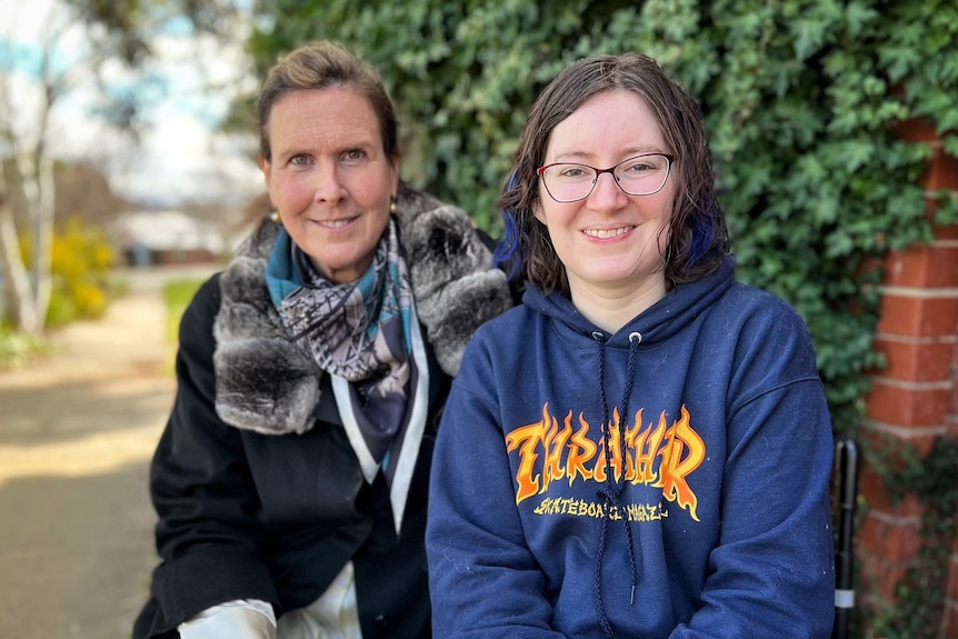 Two women look at the camera, in front of shrubbery.