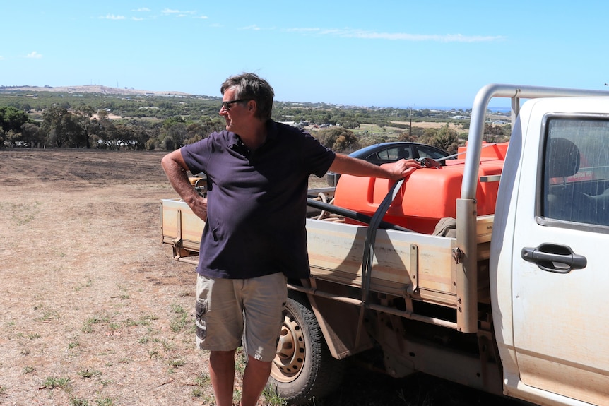 Man looking off to left in sunglasses, tshirt and shorts, leaning on a ute with red tanks on back for firefighting.