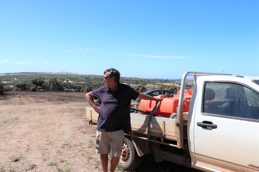 A man stands looking out at paddocks with one arm resting the back of his ute
