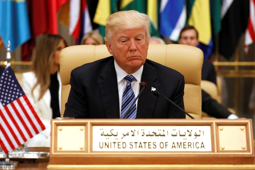 US President Donald Trump looking down while seated in front of row of flags of Middle Easter countries