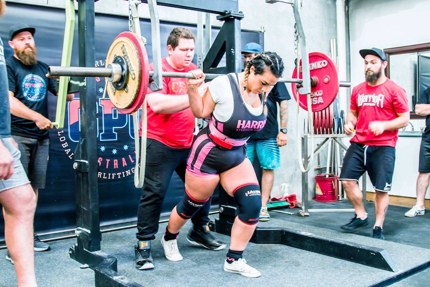 A woman wearing gym shorts, knee braces and waist support lifts a heavy barbell at a gym surrounded by a team of supporters.