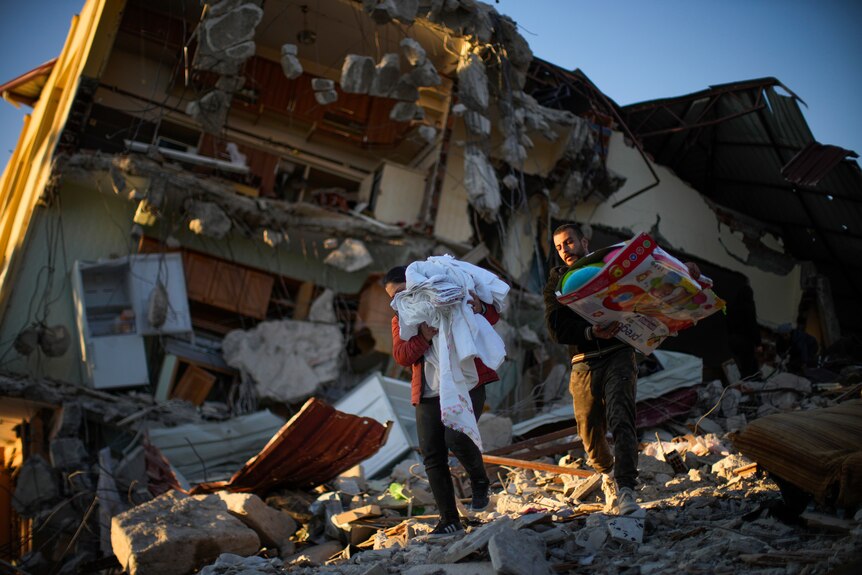 People carry items in their arms in front of a destroyed building. 
