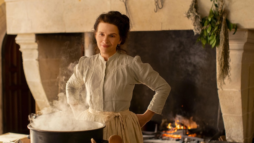 Juliette Binoche in old style clothing, stands in a kitchen with an open fire, in front of a steaming pot
