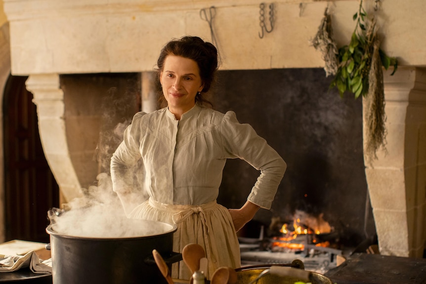 Juliette Binoche in old style clothing, stands in a kitchen with an open fire, in front of a steaming pot