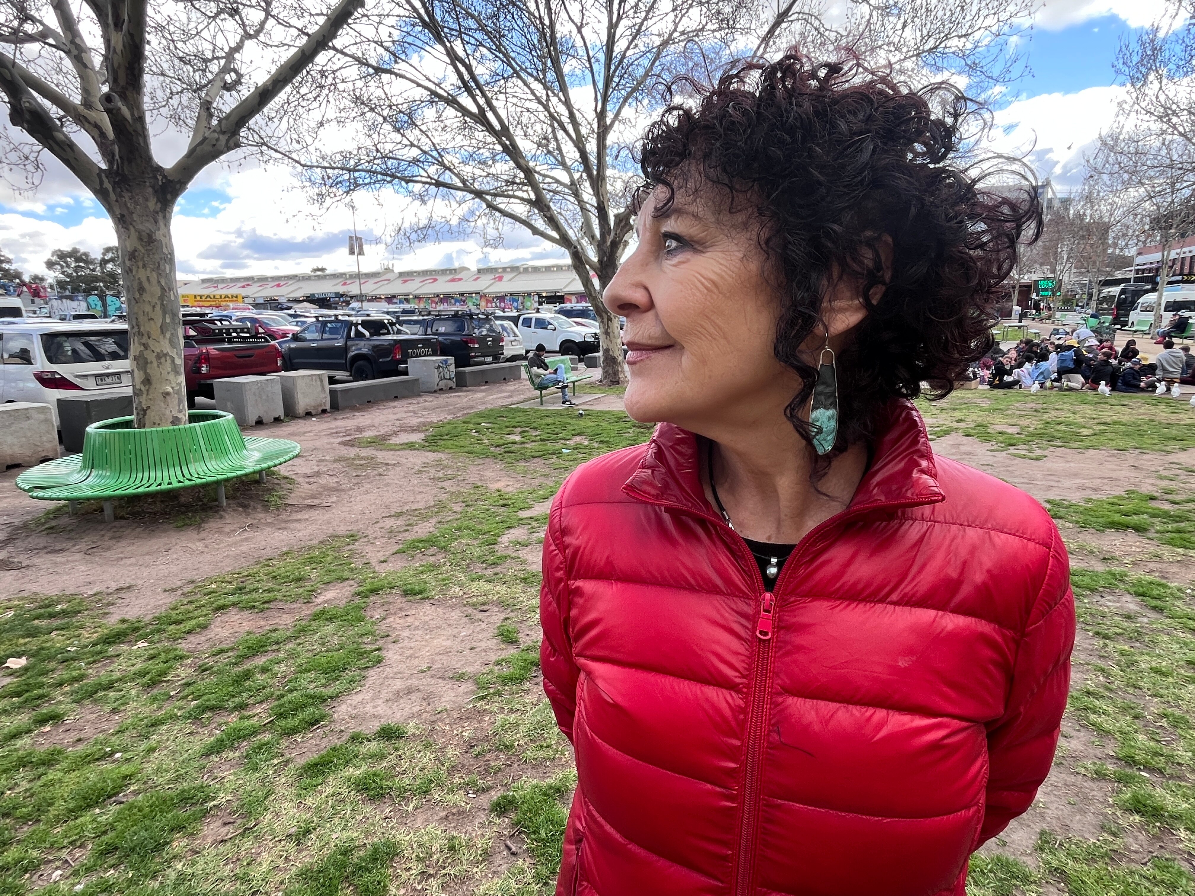 Woman with curly hair and a red jacket standing outside a market looking towards an open-air car park.