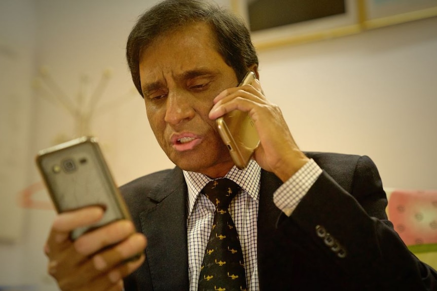 Abul Kalam holds a phone to his ear while also looking at a mobile phone he is holding with his other hand.