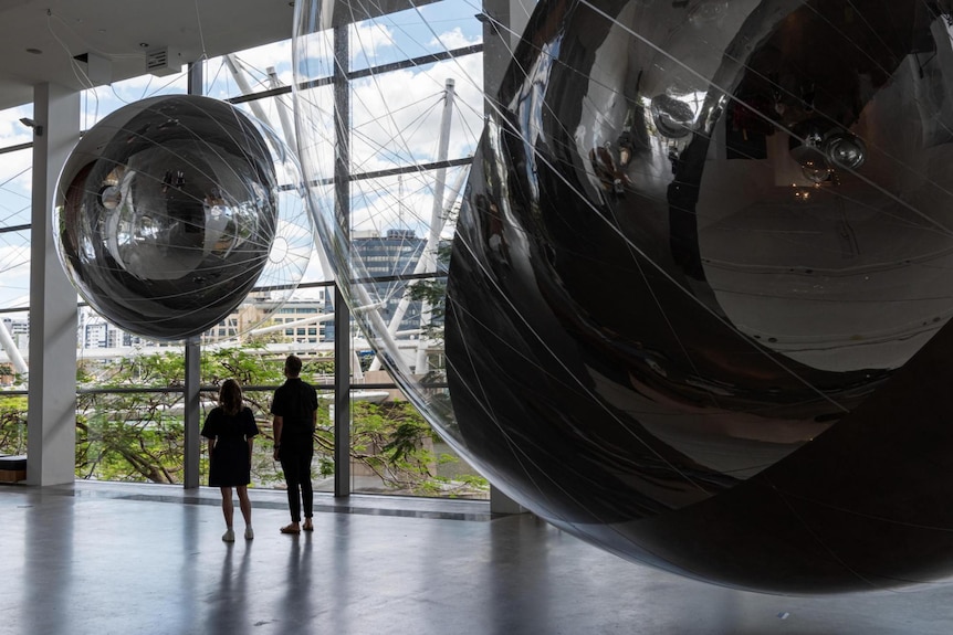 A man and a woman look up at a large silver sphere sculpture. Another silver sphere can be seen in the foreground.