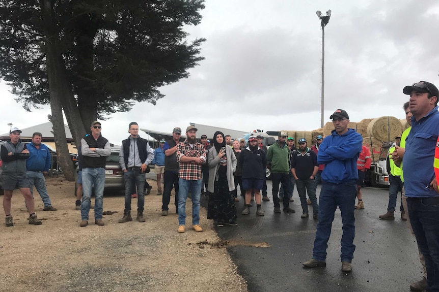 A group of men and women in work clothes assembled near trucks loaded with hay on an overcast-looking day.