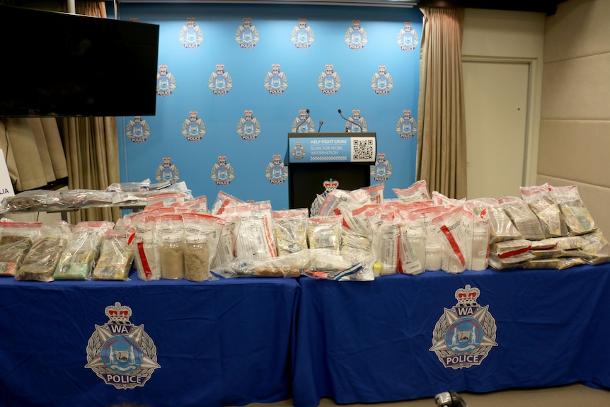 The drugs and cash sit in plastic bags on a table in front of a WA Police logo-covered wall.