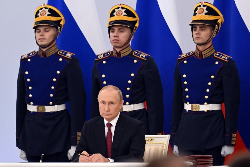 Vladimir Putin siede a una scrivania con in mano una penna, dietro di lui tre ufficiali militari in alta uniforme