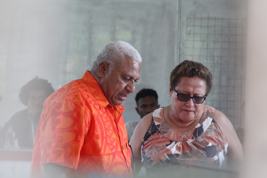 A Fijian man wearing a bright orange shirt stands behind a woman wearing sunglasses in a polling centre