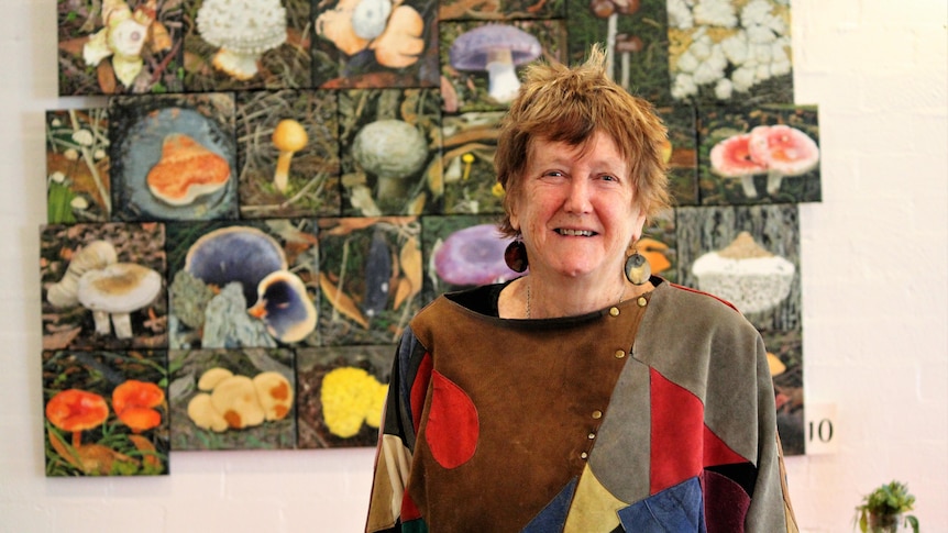 Smiling older woman, short red hair, wears bright, large patterned jumper, stands in front of colourful fungi paintings.