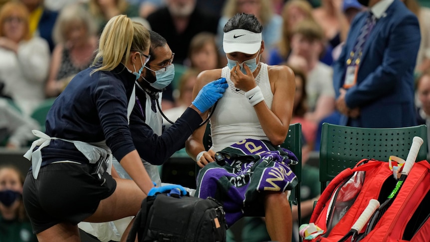 A distressed female tennis player wearing a mask sits at courtside as a doctor uses a stethoscope to check her breathing. 