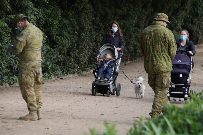 Women wearing masks pushing prams as Defence Force members watch.