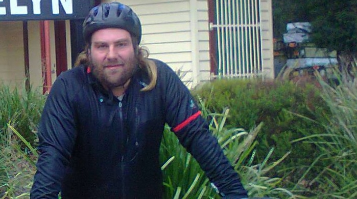 A bearded man stands next to a bicycle.