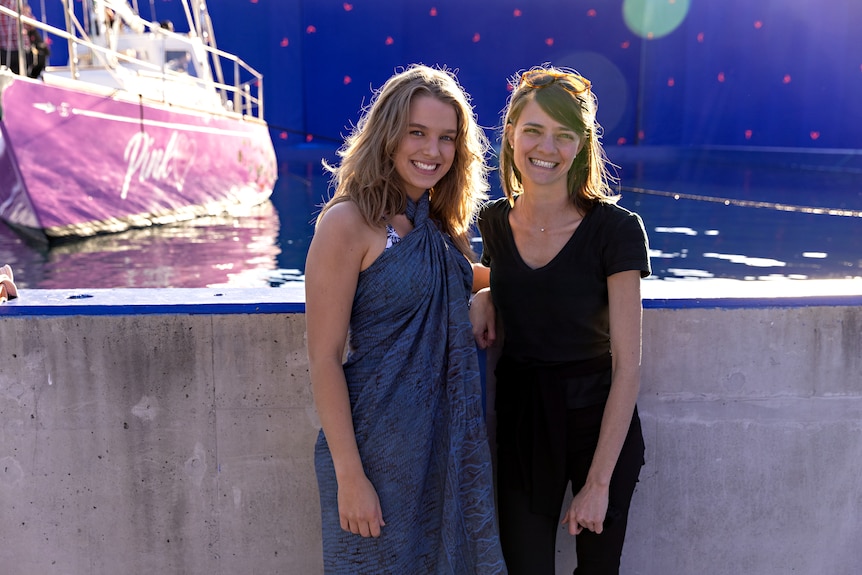 Tegan and Jessica smile for a photo on set with a pink sailing ship docked in front of a blue green screen behind them.
