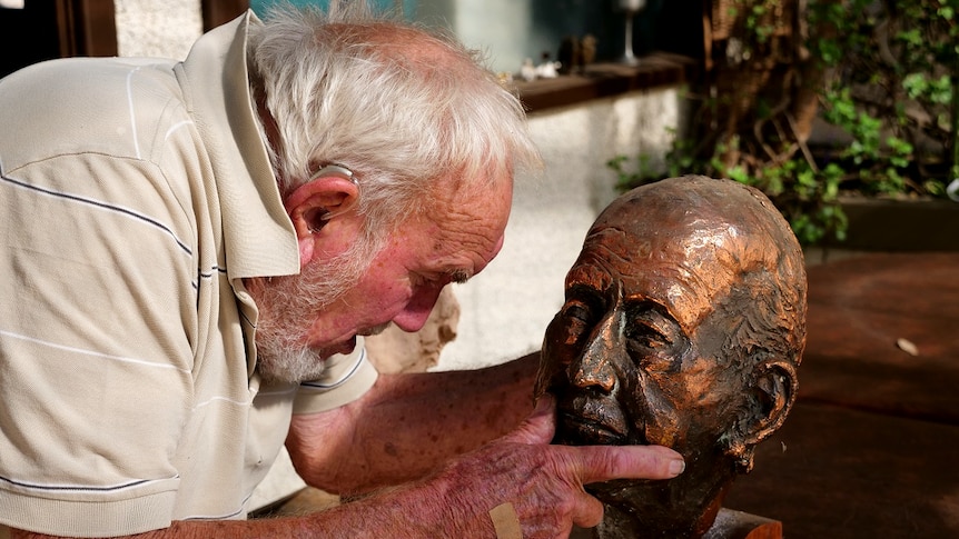 An older artist leans down to touch a bronzed scupture.